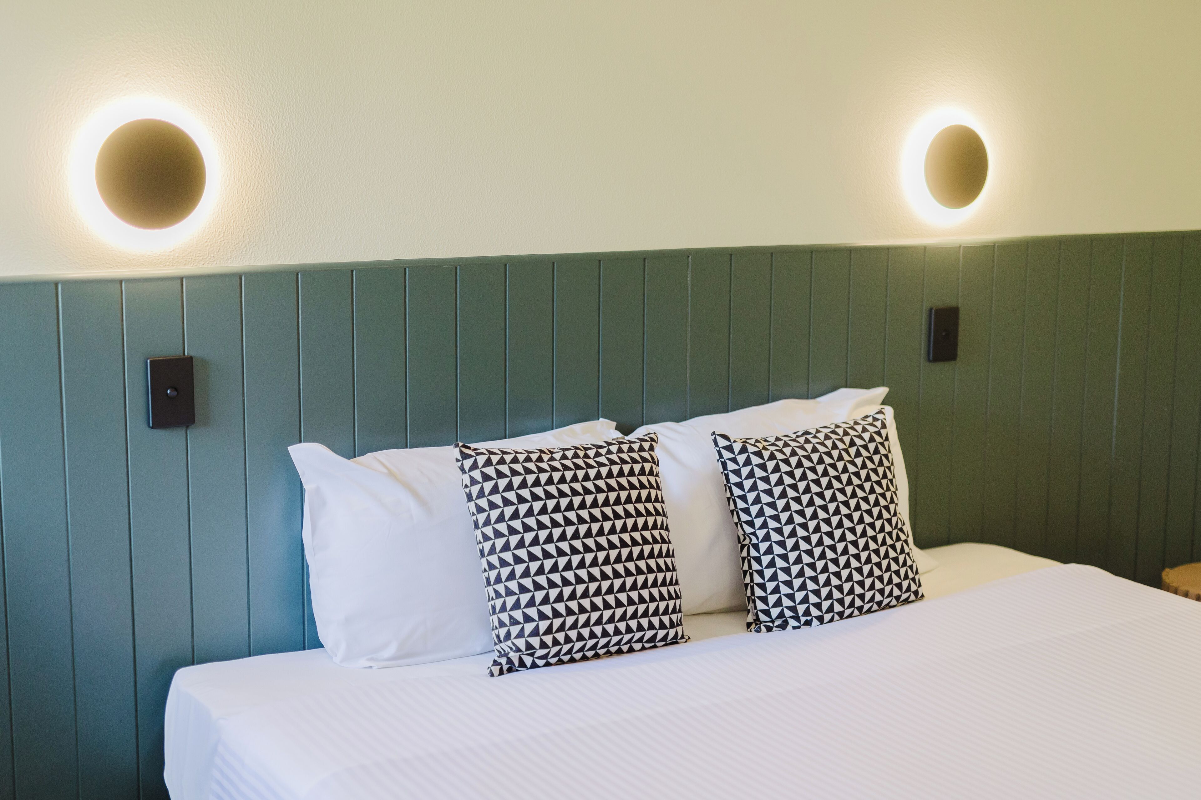 A neatly made bed with white bedding and patterned pillows, a green paneled headboard, and two wall-mounted light fixtures.
