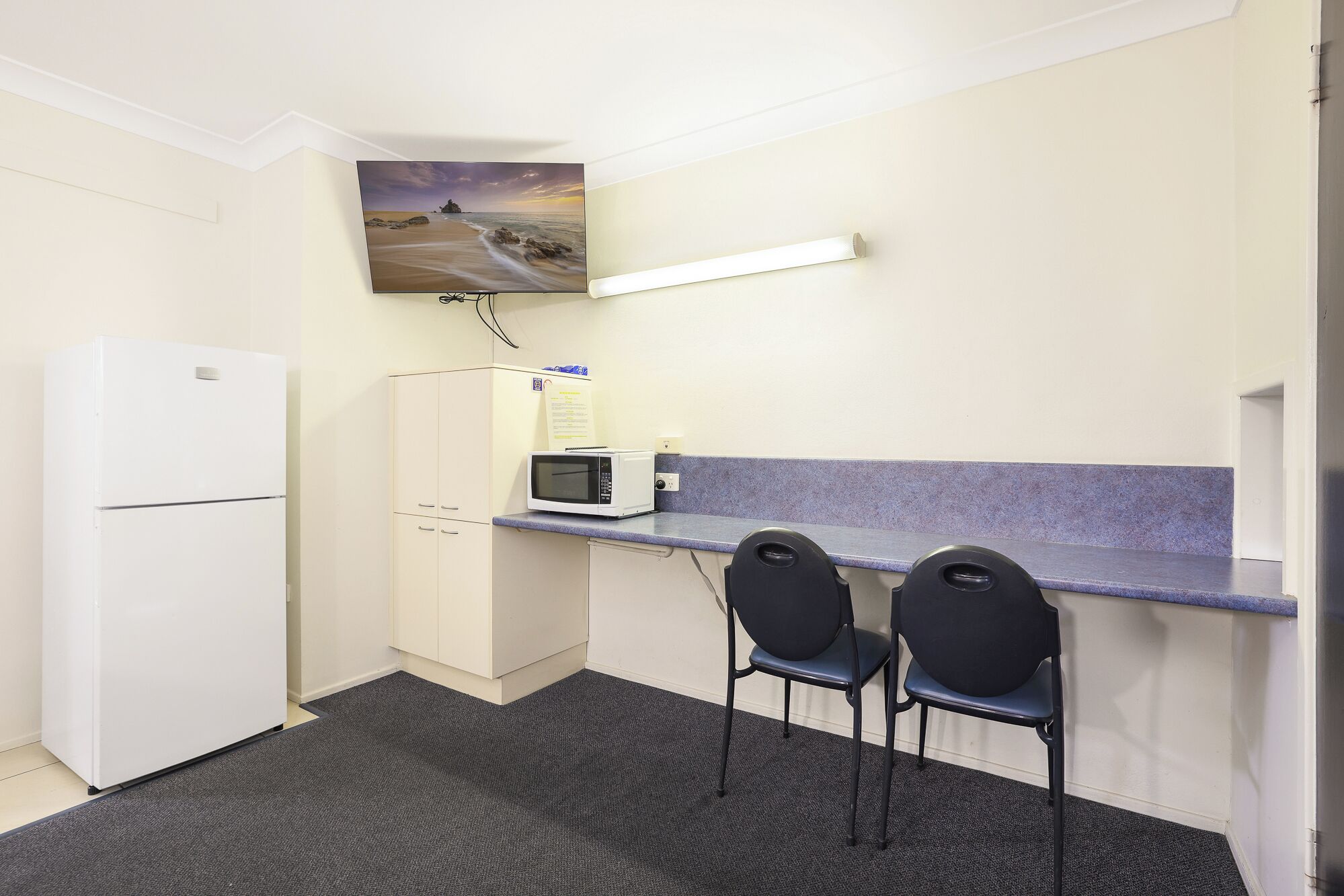 A small kitchenette with a refrigerator, microwave, and a sink. There are two chairs and a painting of a beach scene on the wall.