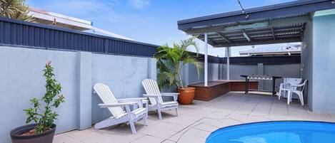 A backyard with a pool, two white lounge chairs, a barbecue grill, and a potted plant.