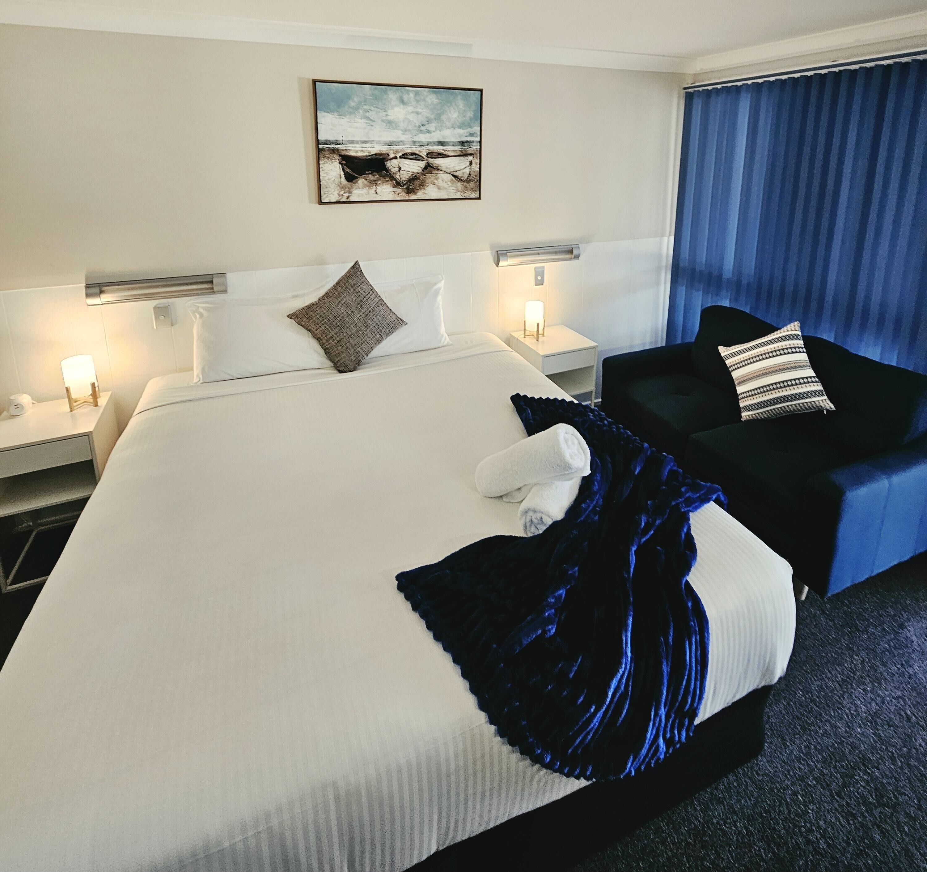 A hotel room with a large bed, two bedside tables, a nightstand, a chair, a blanket, and a painting on the wall.