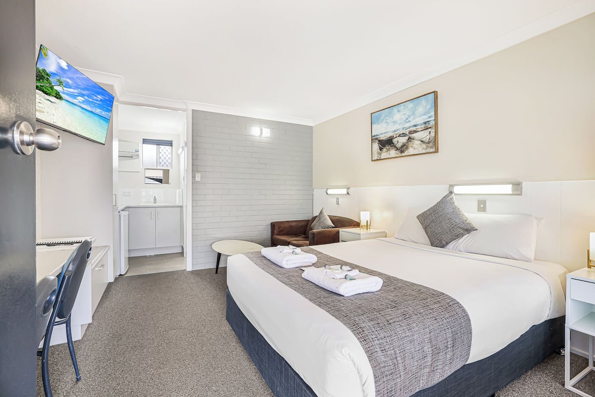 Habitación de hotel con cama, escritorio, silla, mesita, televisión y baño visible a través de una puerta abierta.