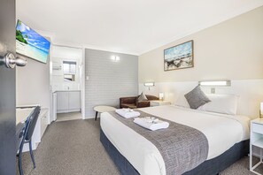 A hotel room with a bed, a desk, a chair, a small table, a TV, and a bathroom visible through an open door.
