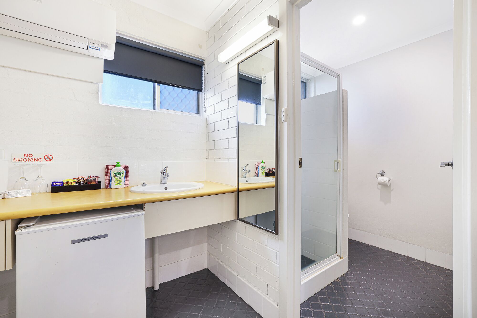A bathroom with a white tiled wall, a yellow countertop, a sink, a mirror, a glass shower enclosure, and a small refrigerator.