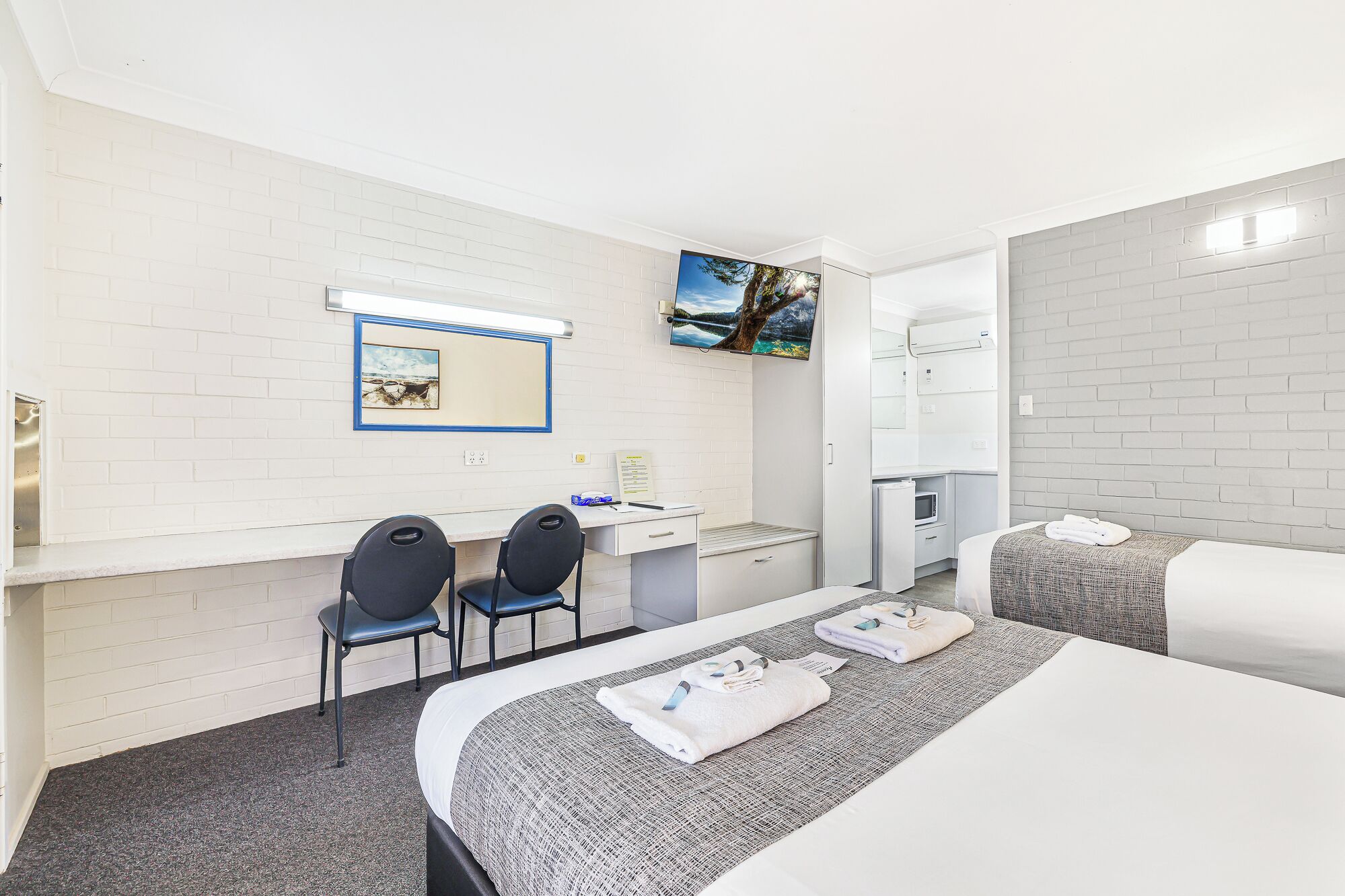 A hotel room with a bed, a desk with two chairs, a flat-screen TV mounted on the wall, and a large mirror.