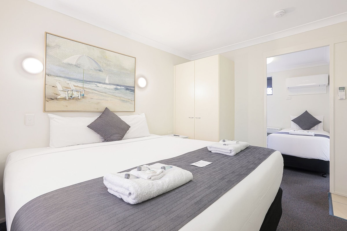 Habitación de hotel con una cama grande, una mesita de noche, un espejo y un cuadro de una playa.