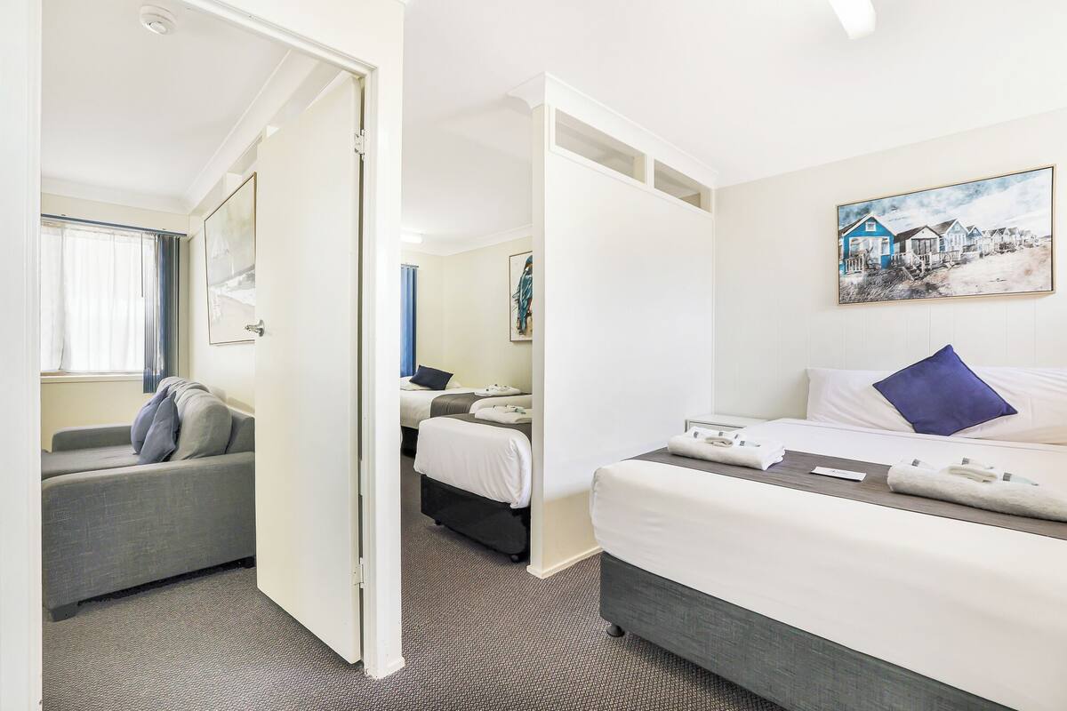 Habitación de hotel con cama, sofá y escritorio. Se observa un cuadro en la pared y una ventana con cortinas.