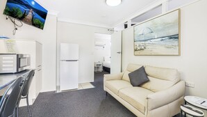 Kompakti hotellihuone, jossa on pieni keittiönurkkaus, sohva, työpöytä ja taulu, jossa on kuvattuna rantamaisema.