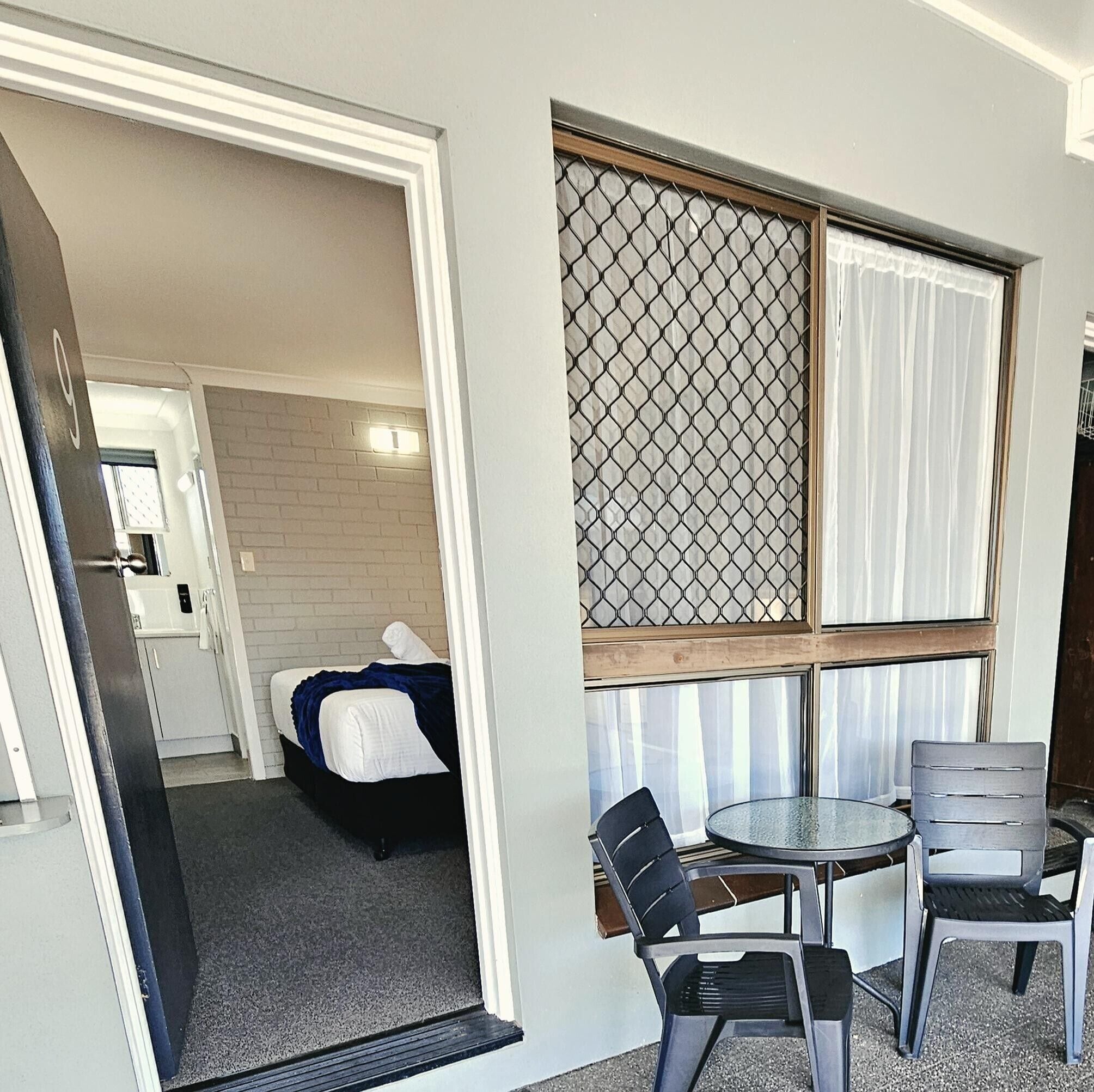 A room with a bed, a small table with chairs, a window with curtains, and a door leading to another room.
