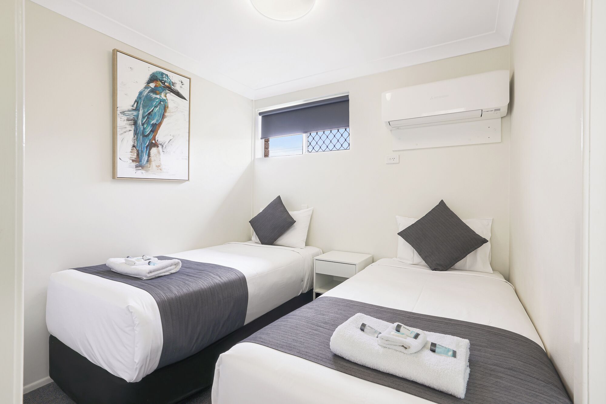 A hotel room with two beds, a painting of a bird, an air conditioning unit, and a window with a view.