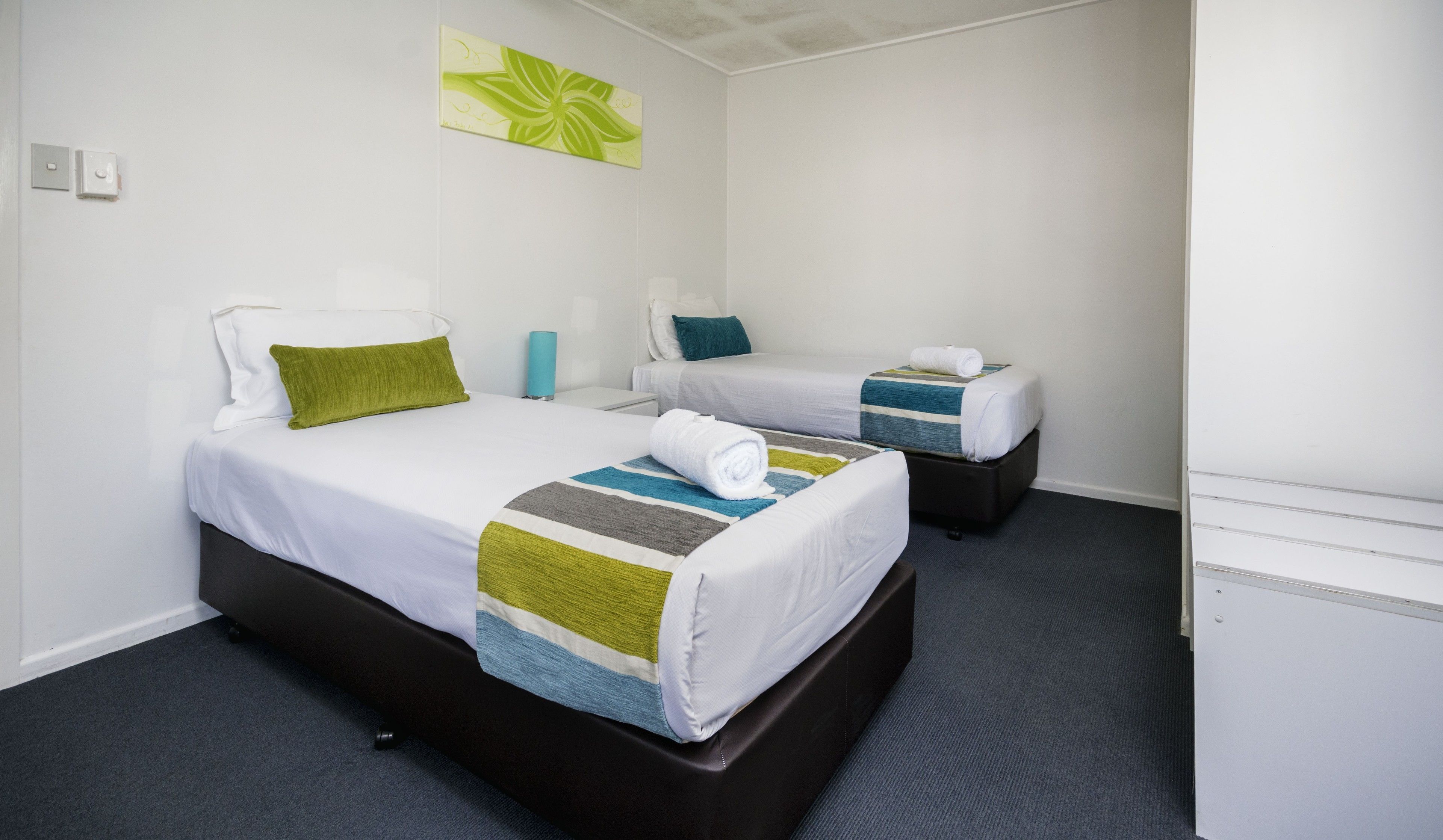 Hotellihuone, jossa on kaksi sänkyä. Kummassakin sängyssä on valkoinen patja, vihreä- ja siniraidallinen peitto, ja toisessa sängyssä on rullattu valkoinen pyyhe.