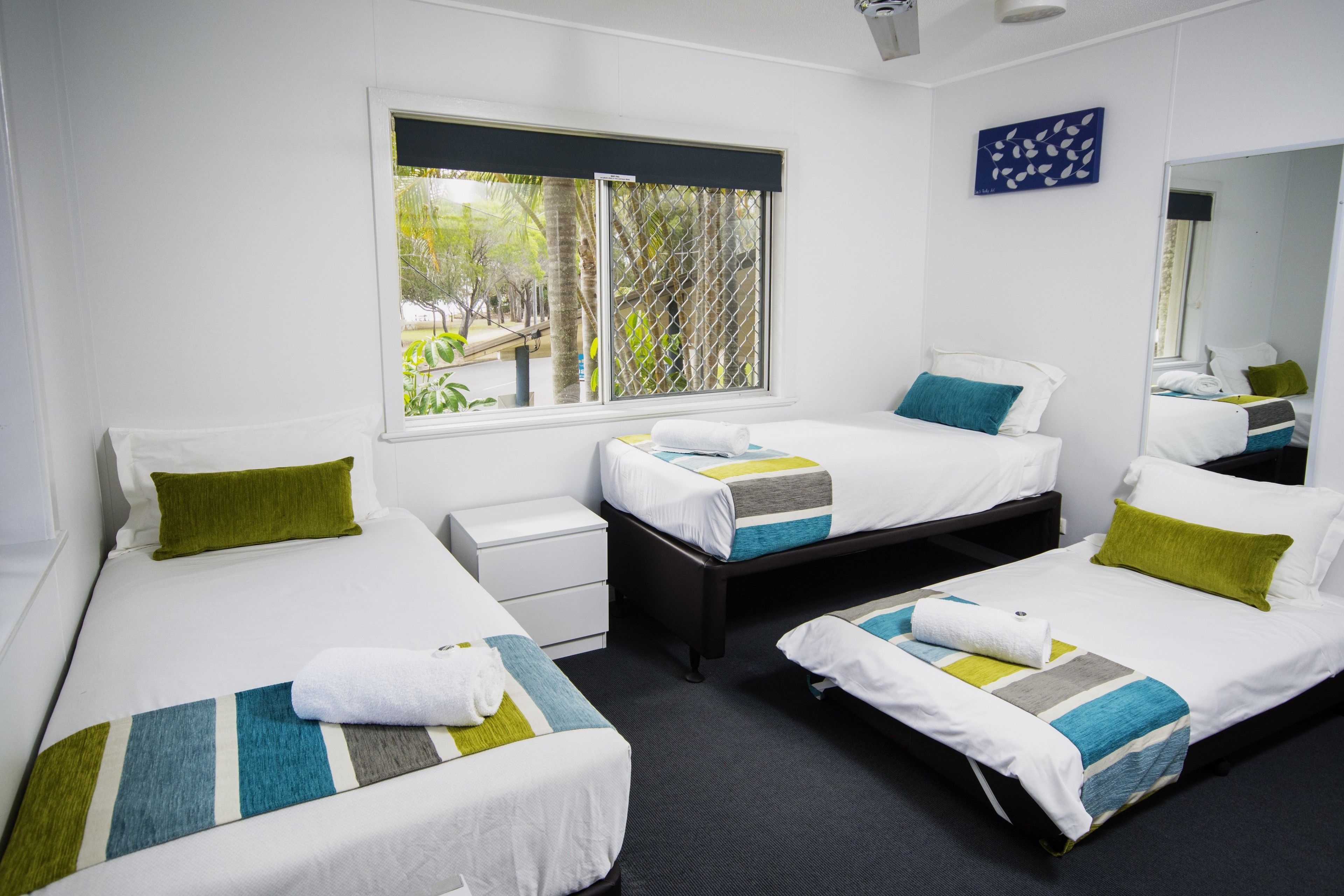 A hotel room with two single beds, a small bedside table, a large window with a view of greenery, and a mirror on the wall.