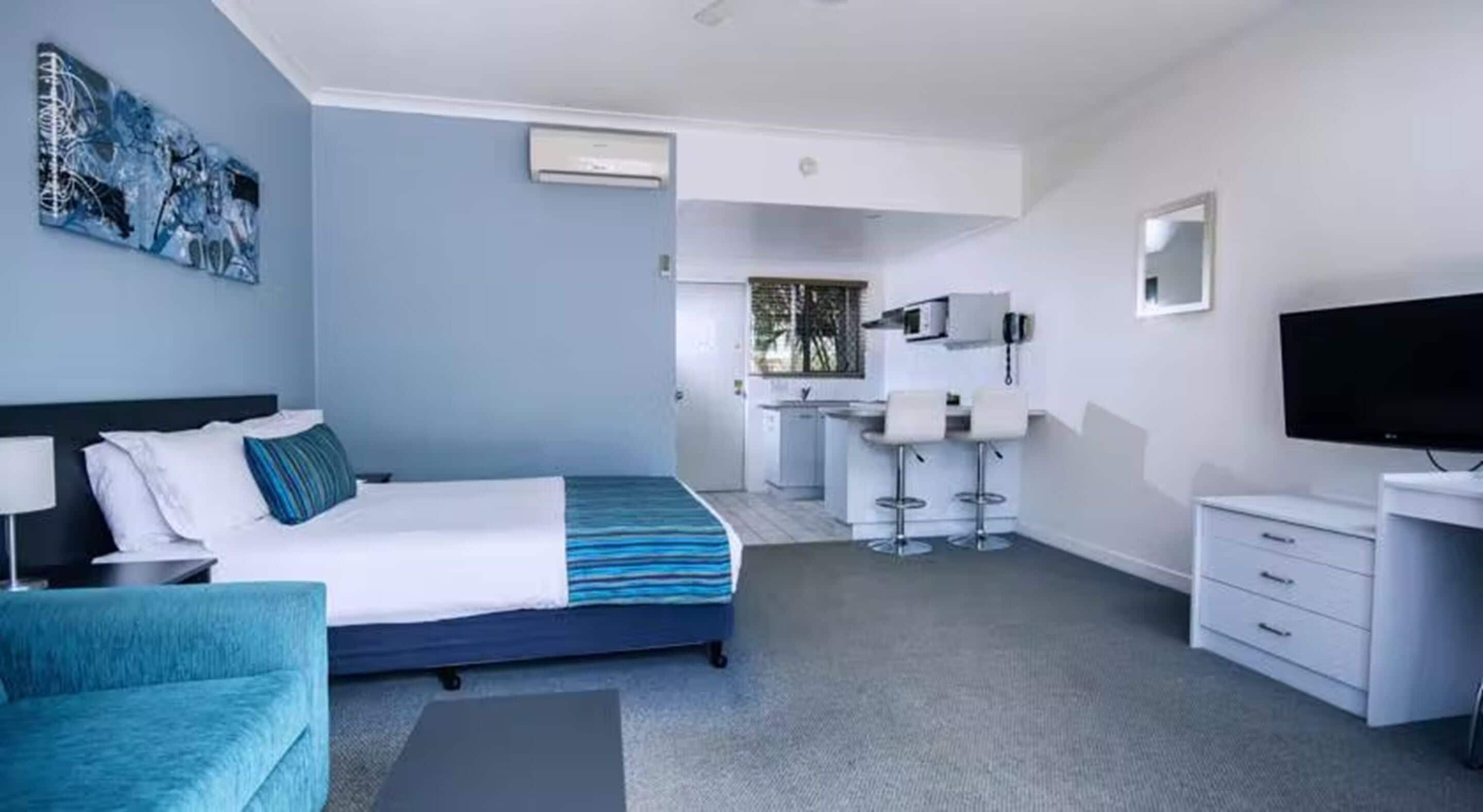 A modern hotel room with a bed, a desk, a TV, and a kitchenette.