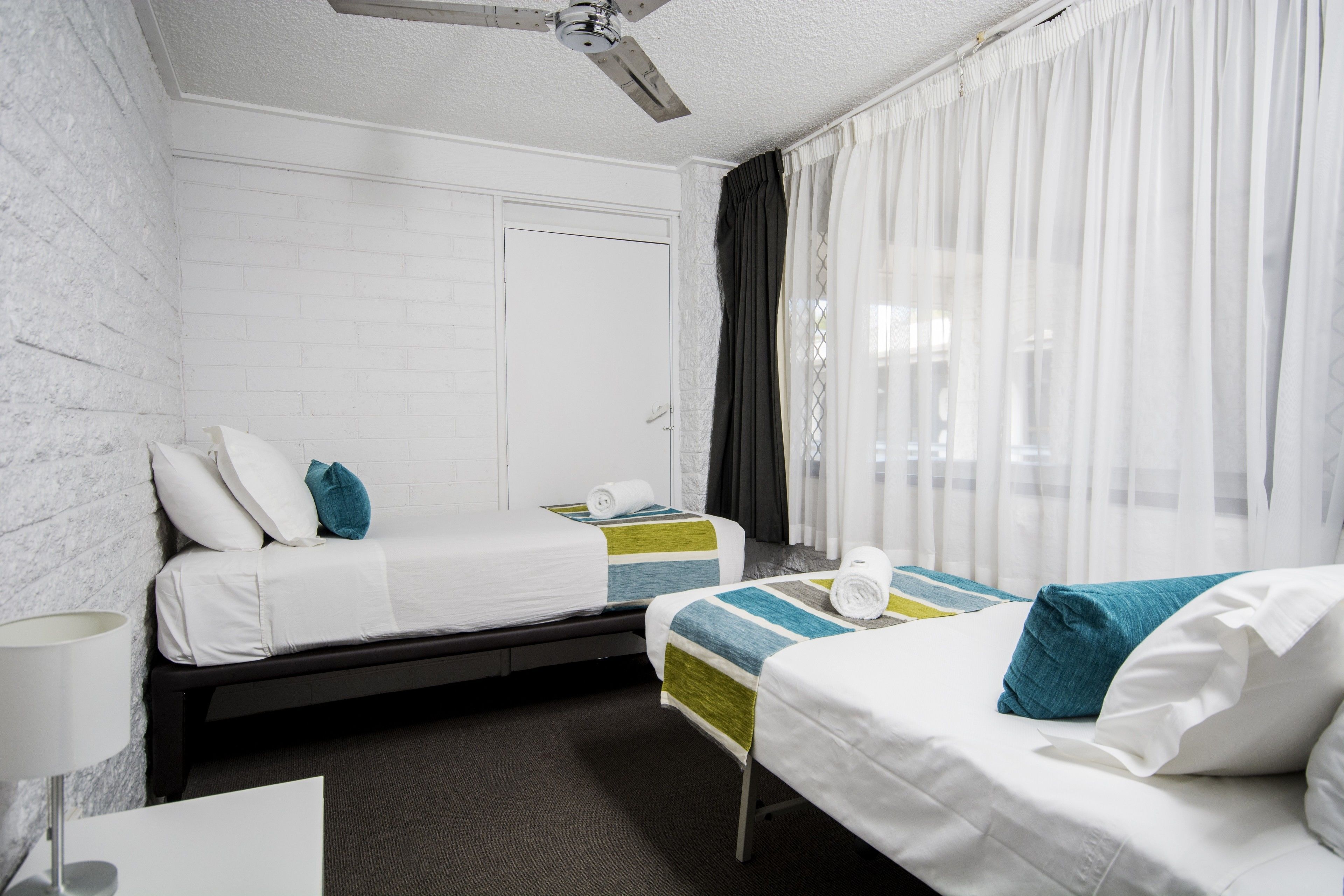 Habitación de hotel con dos camas, ventilador de techo, ventana con cortinas traslúcidas y pared de ladrillo blanco.