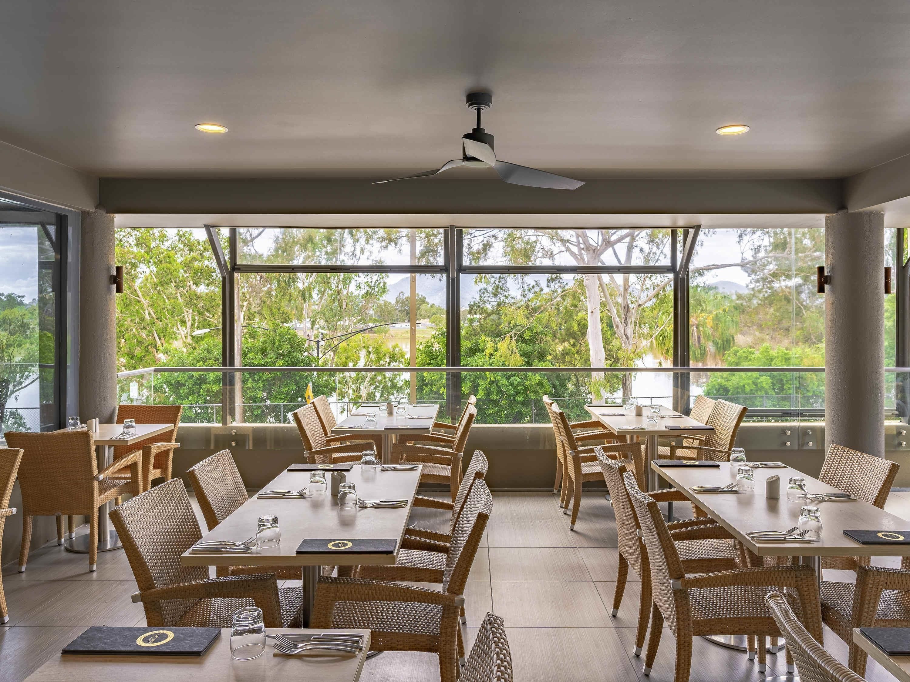 Comedor con mesas y sillas, ventanas amplias con vista a árboles y ventiladores de techo.