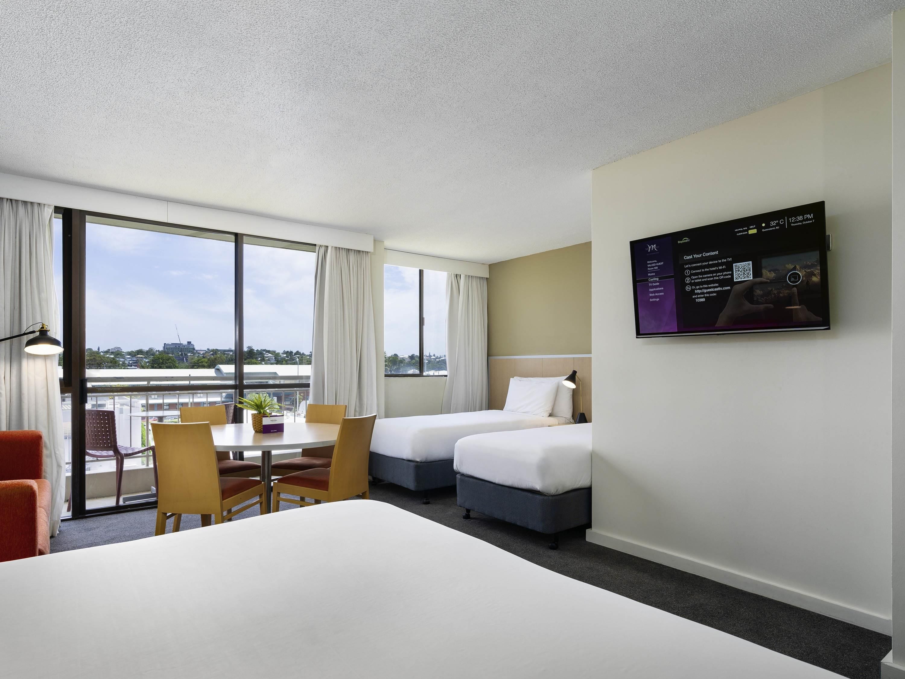 A hotel room with two beds, a desk, chairs, and a large window with a city view.