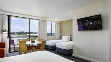 A hotel room with two beds, a desk, chairs, and a large window with a city view.