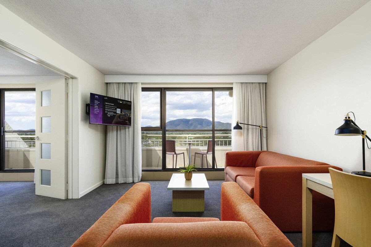 Habitación de hotel con sofá, una mesita, escritorio, televisor y balcón con vistas a la montaña.