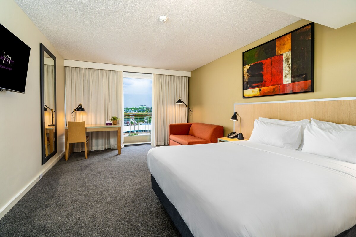 A hotel room with a large bed, a desk, a chair, a sofa, a TV, and a painting on the wall.