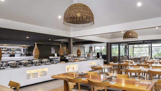 A modern restaurant with wooden tables and chairs, a buffet area, and large windows offering a view of the outdoors. - Mercure Rockhampton (Rockhampton)