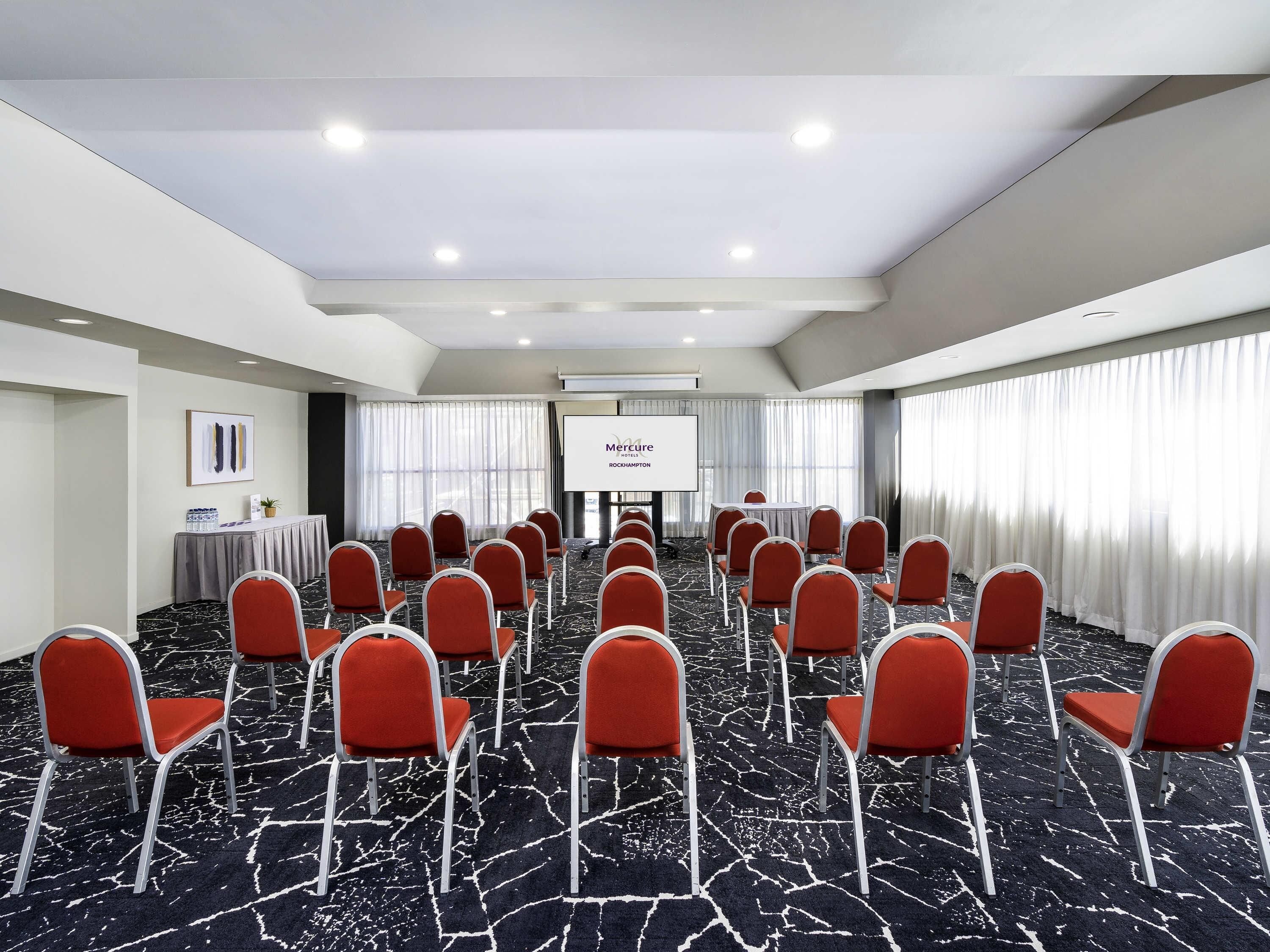 A conference room with rows of red chairs, a projector screen, and a presentation screen.