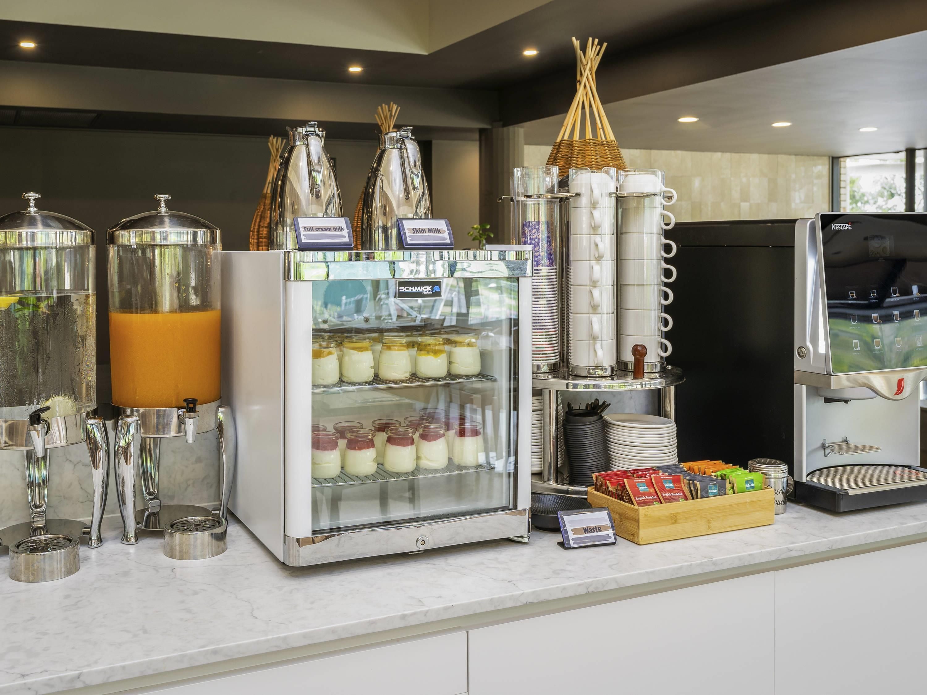 A self-service beverage station with dispensers for full cream and skim milk, a refrigerated display for yogurt, and a coffee machine.