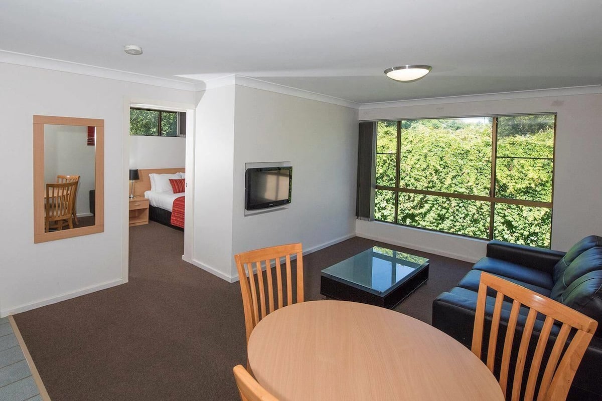 A modern hotel room with a dining area, a sofa, a TV, and a large window with a view of greenery.