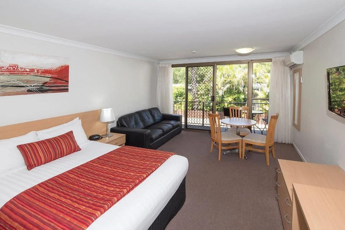 A hotel room with a bed, a black leather sofa, a dining table with chairs, and a desk.
