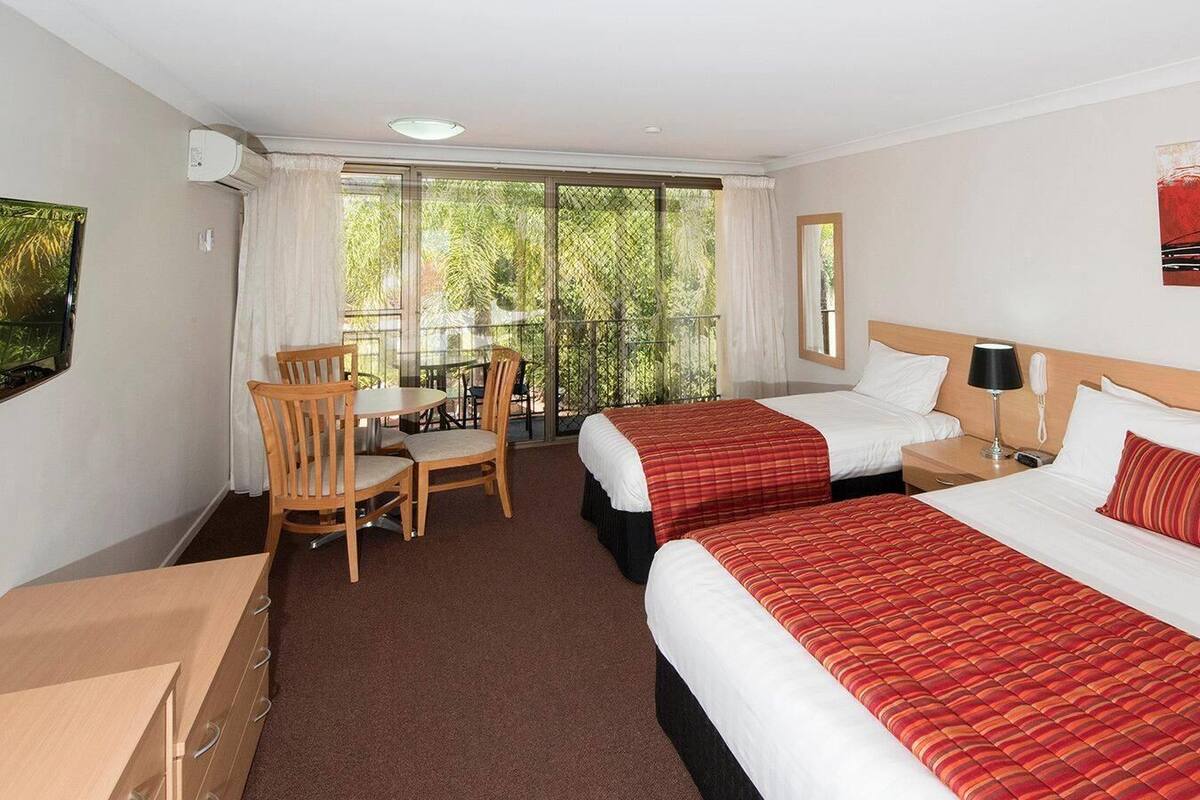A hotel room with two beds, a desk, chairs, and a balcony with a view of trees.