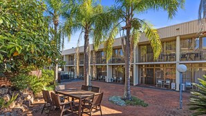 A courtyard with a wooden table and chairs, surrounded by palm trees and a building with balconies. - Comfort Inn Grammar View (East Toowoomba)