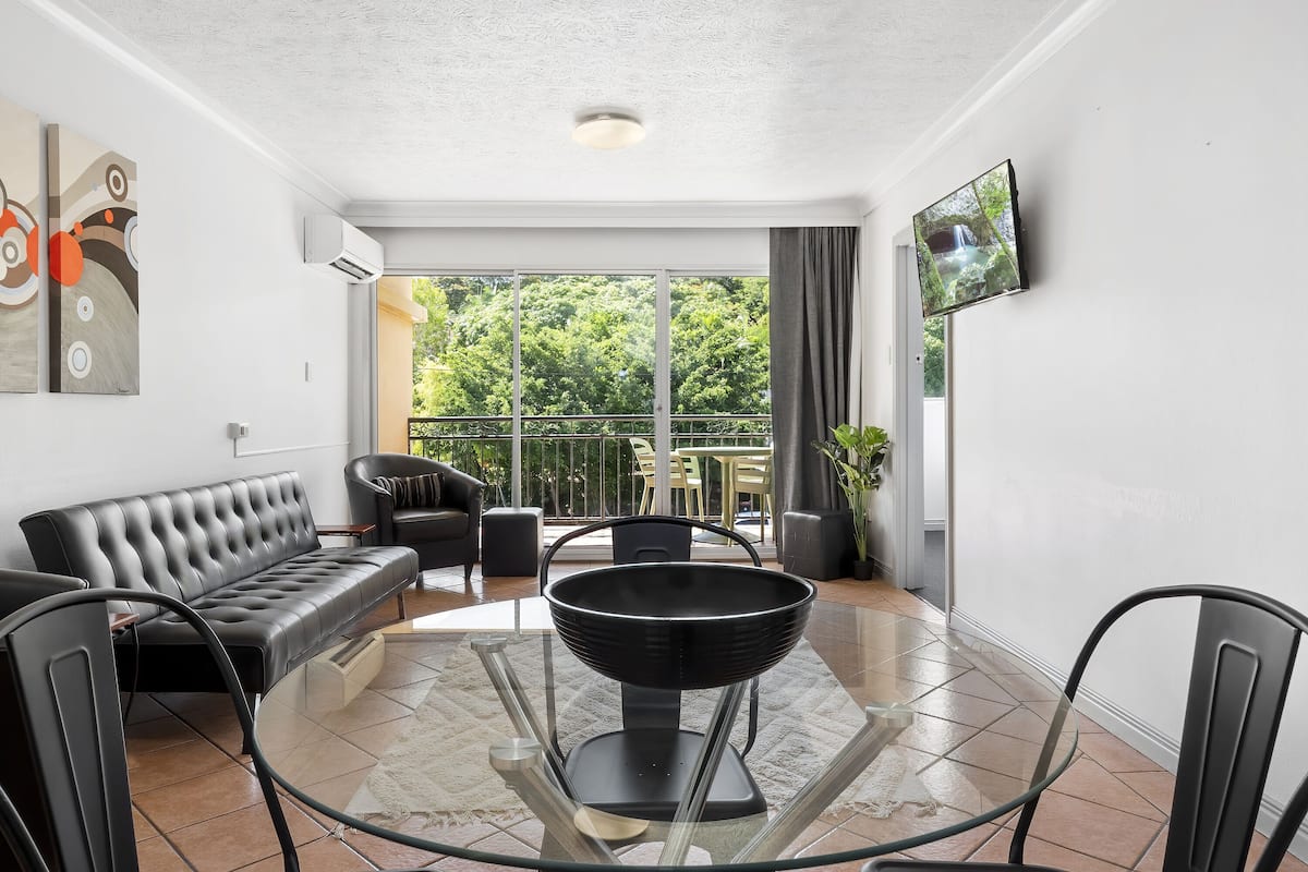 A modern living room with a glass-top coffee table, black leather sofa, and a flat-screen TV mounted on the wall.