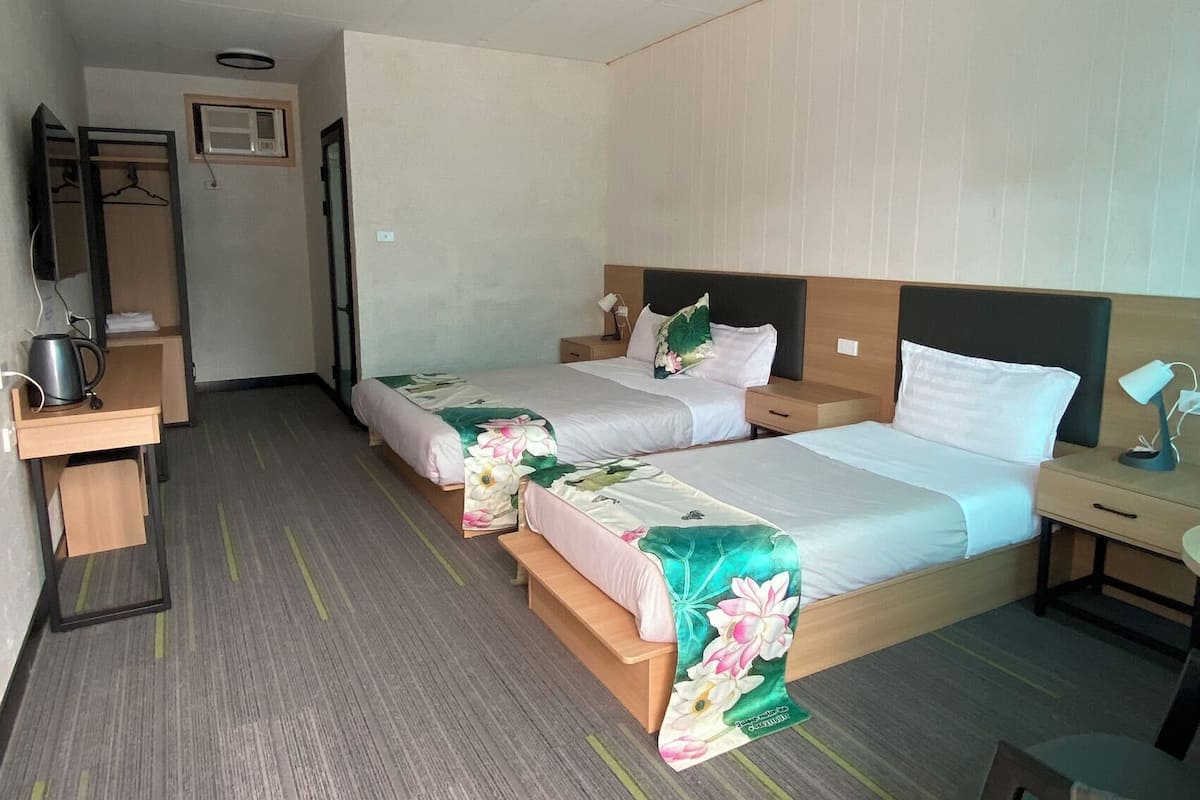 A hotel room with two beds, a desk with a kettle, a chair, and a small table with a lamp.