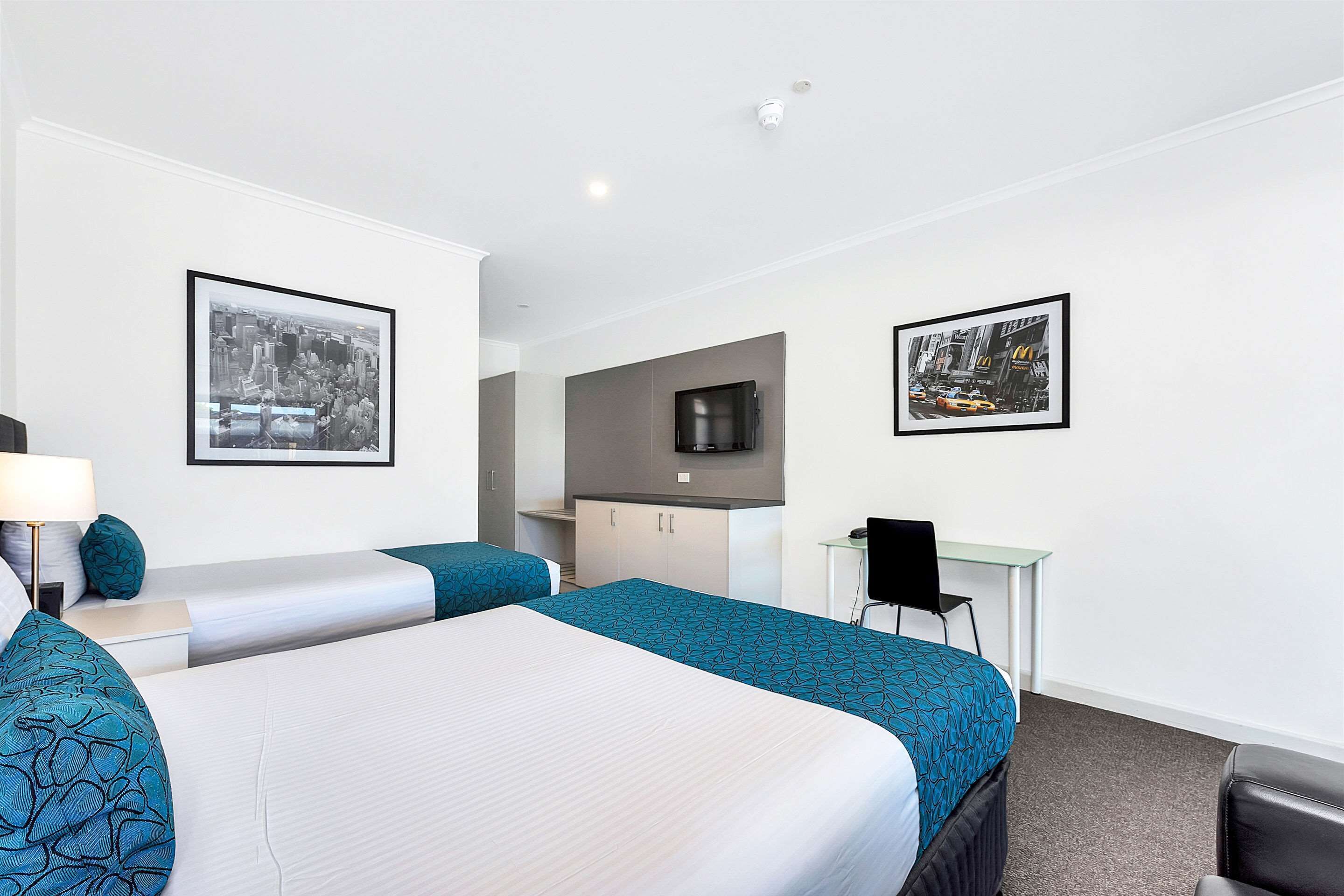 A hotel room with two beds, a desk, a chair, a TV, and framed pictures on the walls.