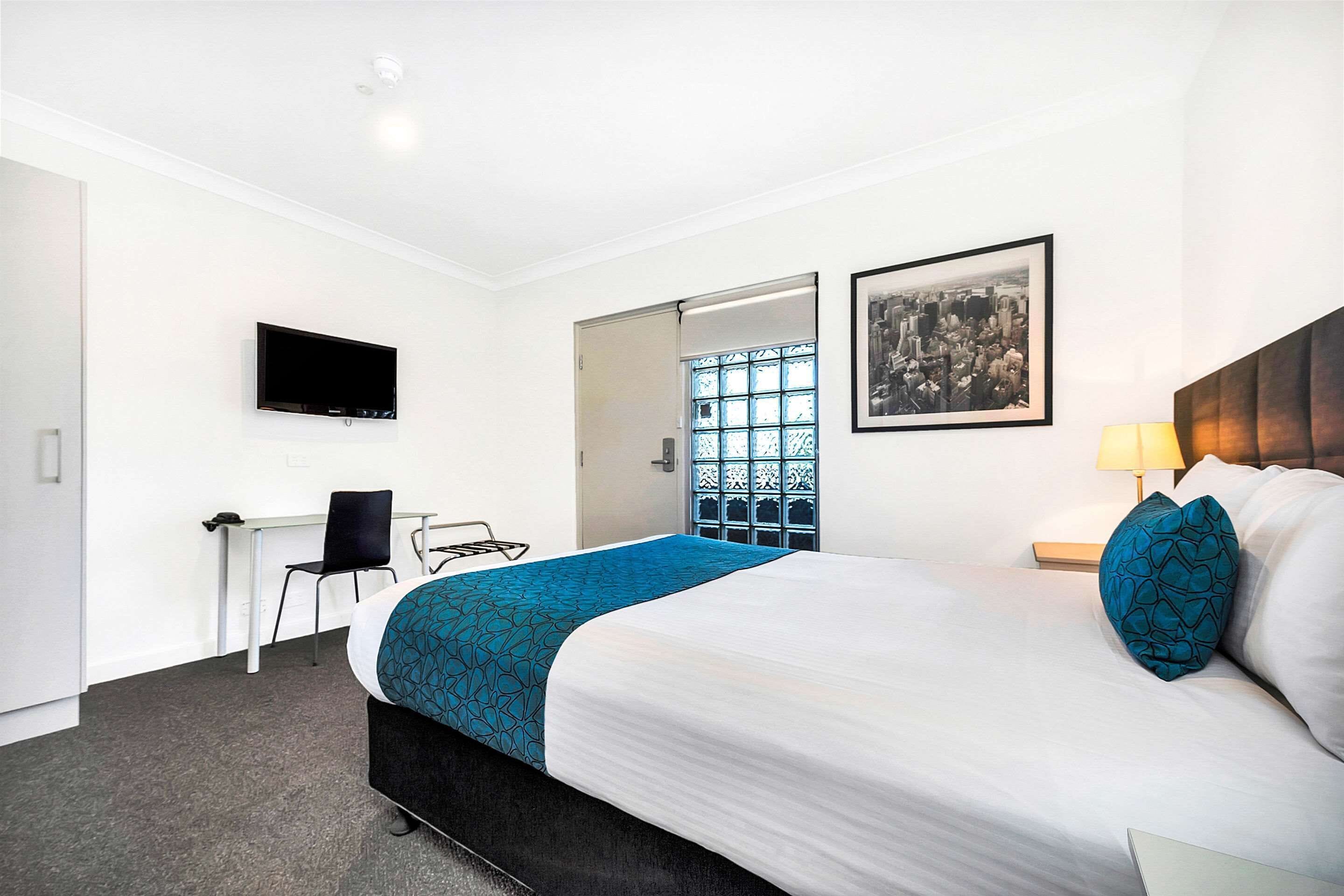 A hotel room with a bed, a desk, a chair, a TV, a window with a view, and a framed picture on the wall.