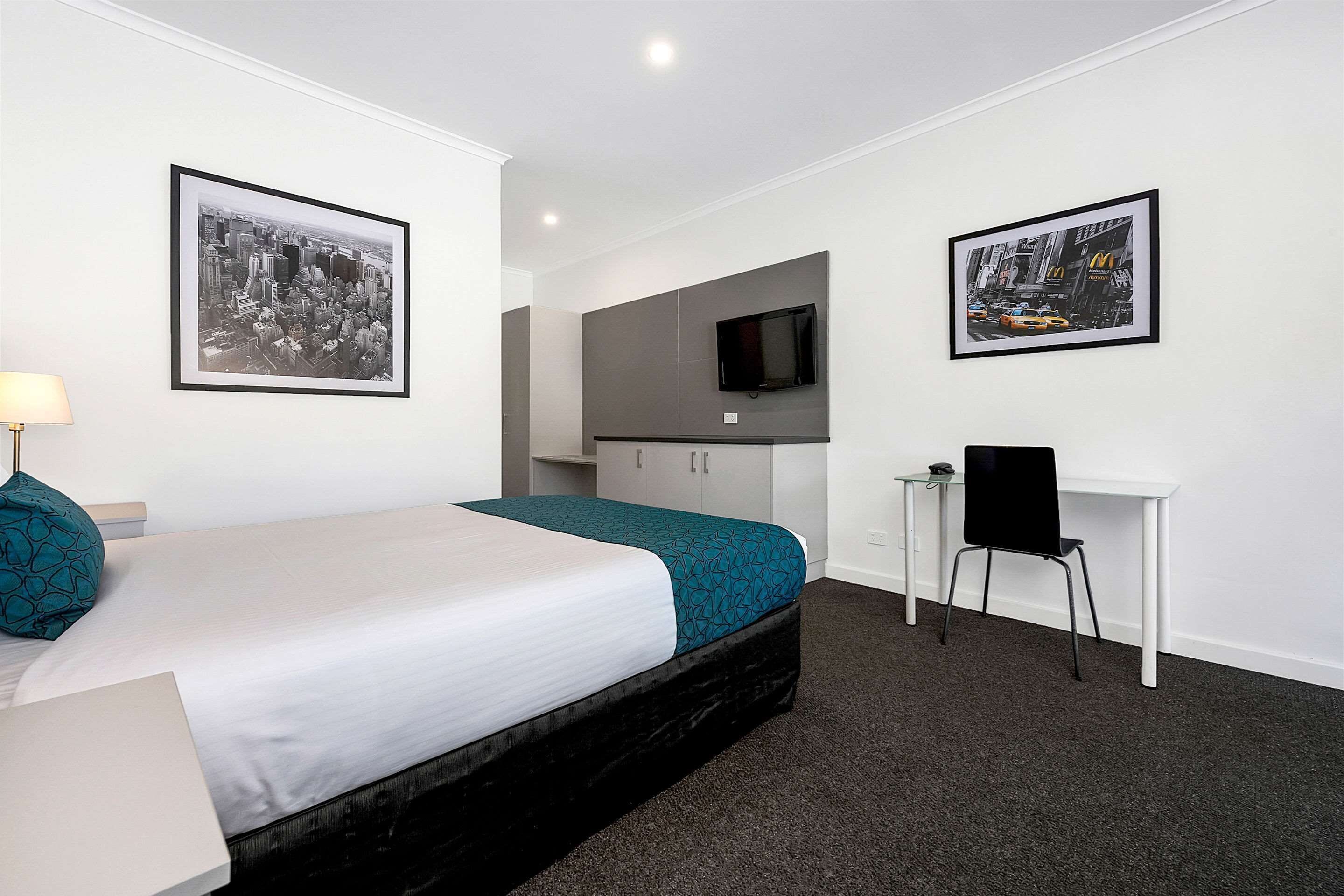 A hotel room with a bed, a desk, a chair, a TV, and two framed pictures on the walls.