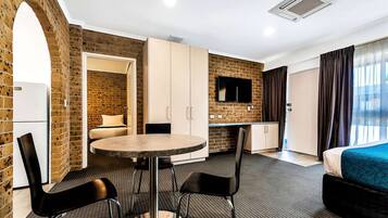 A compact hotel room with a brick wall, a bed, a dining table with chairs, a kitchenette, and a TV.