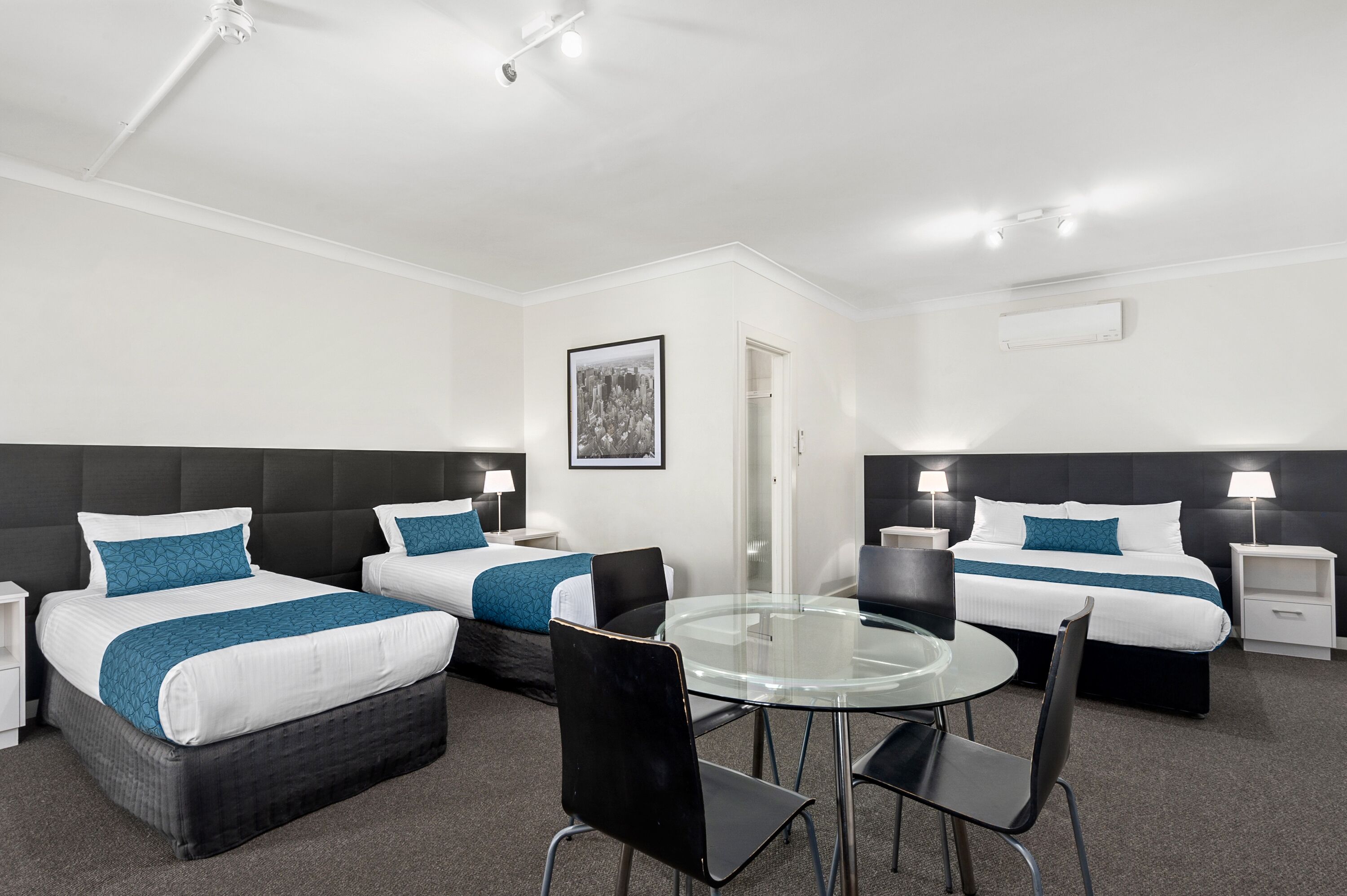 A hotel room with two beds, a glass table, and a chair. There are bedside tables with lamps and a framed picture on the wall.