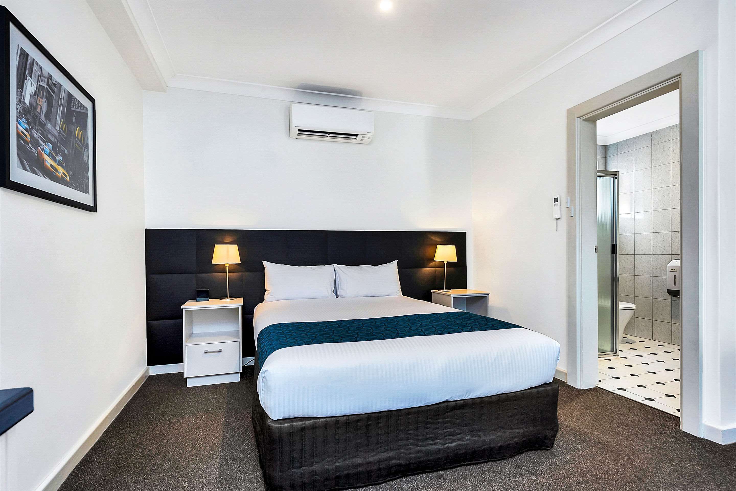 A hotel room with a bed, bedside tables, an air conditioning unit, a framed picture on the wall, and a bathroom visible through an open door.