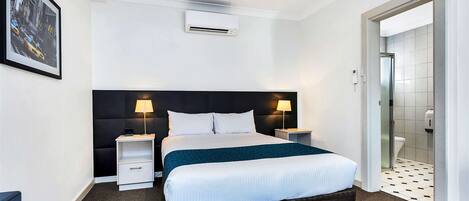 A hotel room with a bed, bedside tables, an air conditioning unit, a framed picture on the wall, and a bathroom visible through an open door.