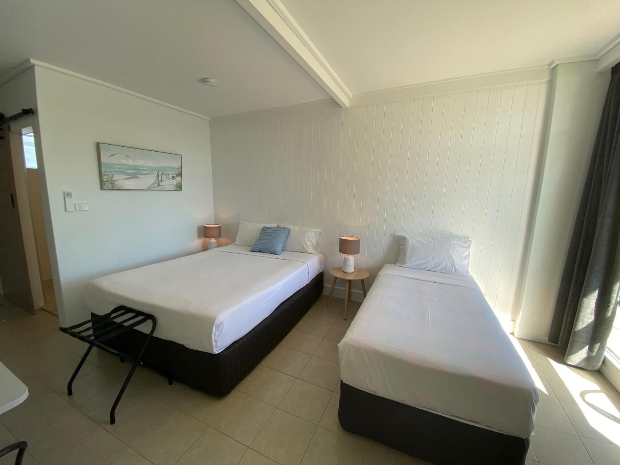 A hotel room with two beds, a small table, a chair, and a painting on the wall.