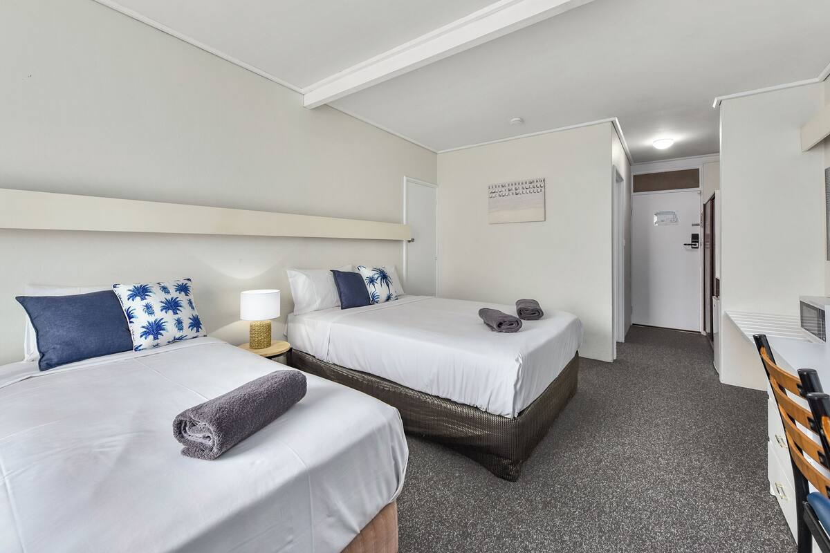 Une chambre d’hôtel avec deux lits, un bureau, une chaise, une lampe et une porte donnant sur une autre pièce.