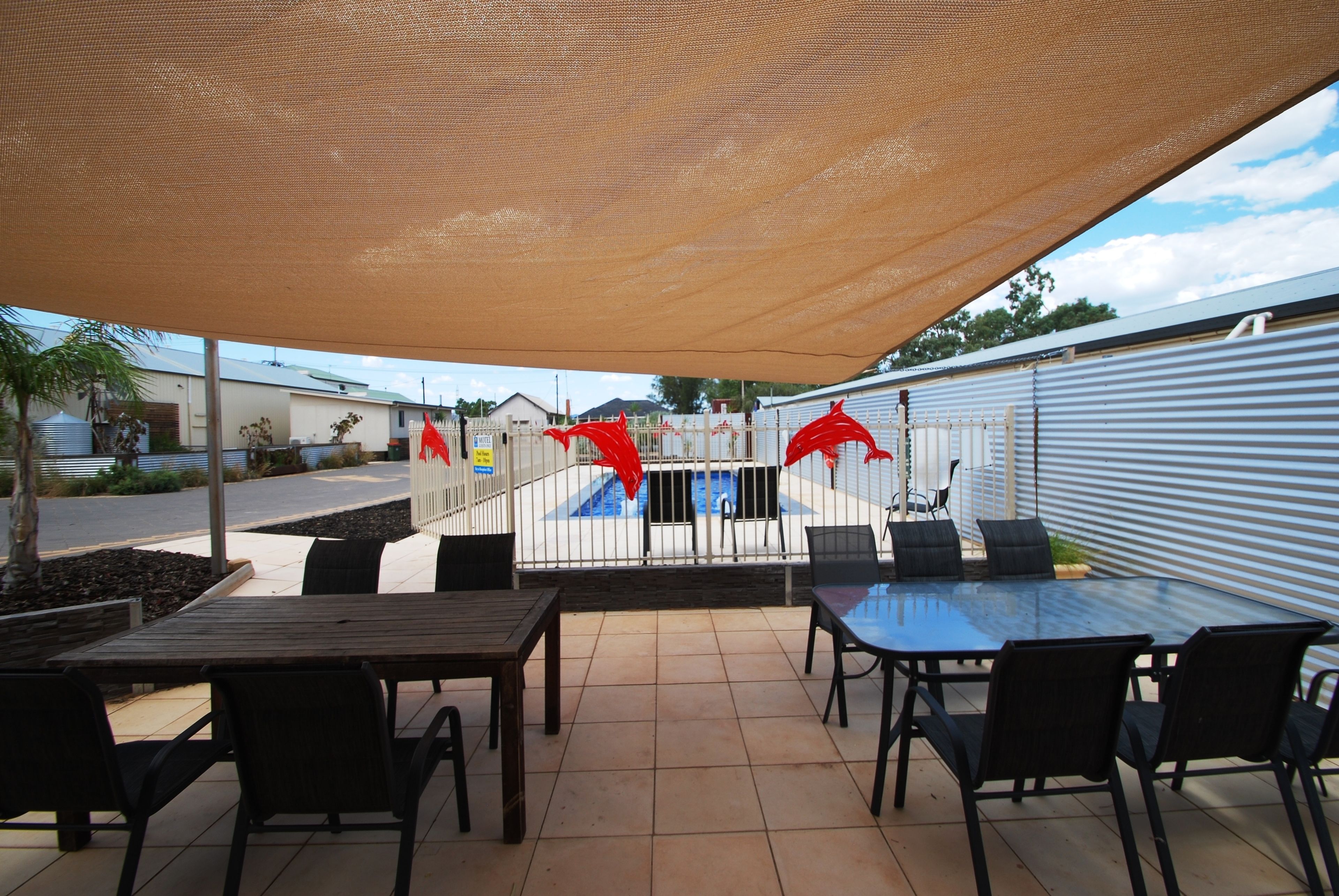 An outdoor patio area with a table and chairs, a pool in the background, and a covered seating area.