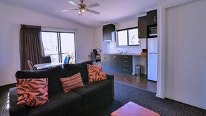 Ein modernes Wohnzimmer mit einer schwarzen Couch, einem orangefarbenen Ottomanen und einem Laptop auf einem Tisch. Im Hintergrund ist eine Küche mit dunklen Schränken und einem Fenster zu sehen.
