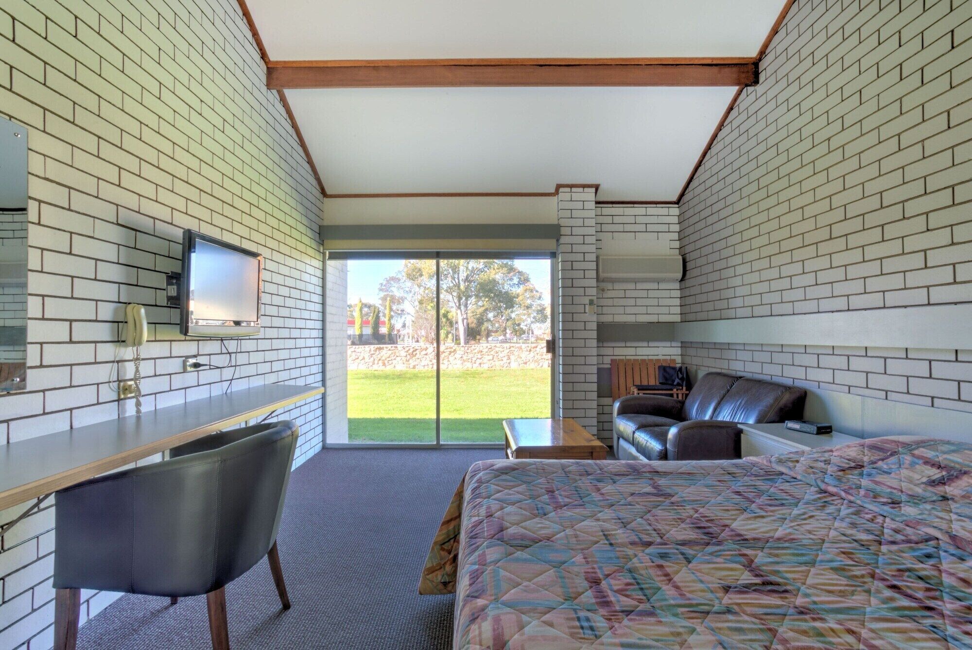 A bedroom with a bed, a desk, a chair, a TV, and a view of a grassy area.
