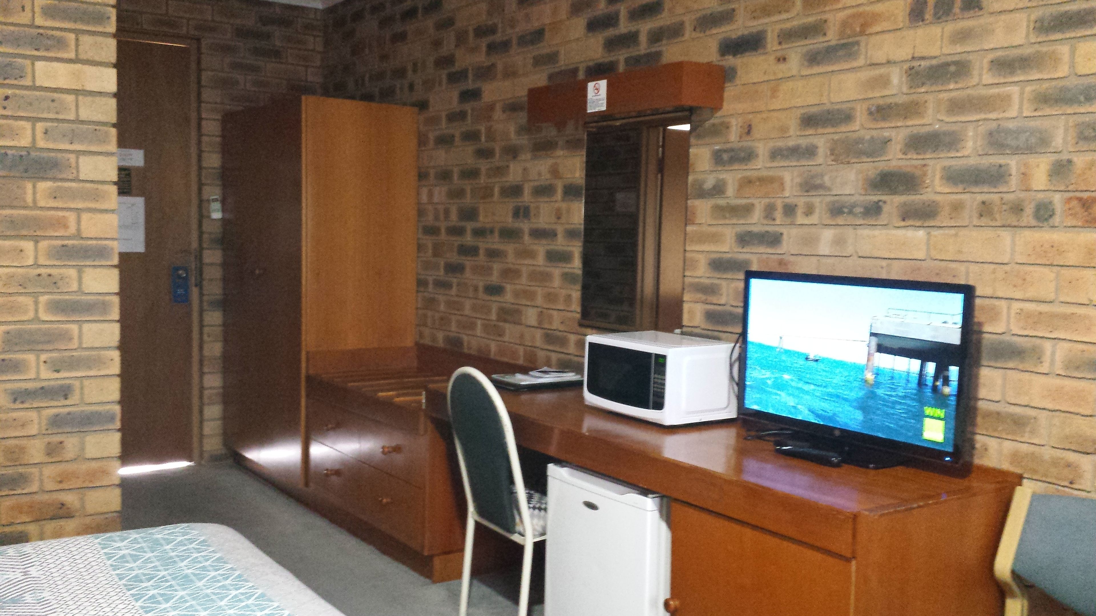 A room with a brick wall, a wooden desk, a microwave, a television, a chair, and a small refrigerator.