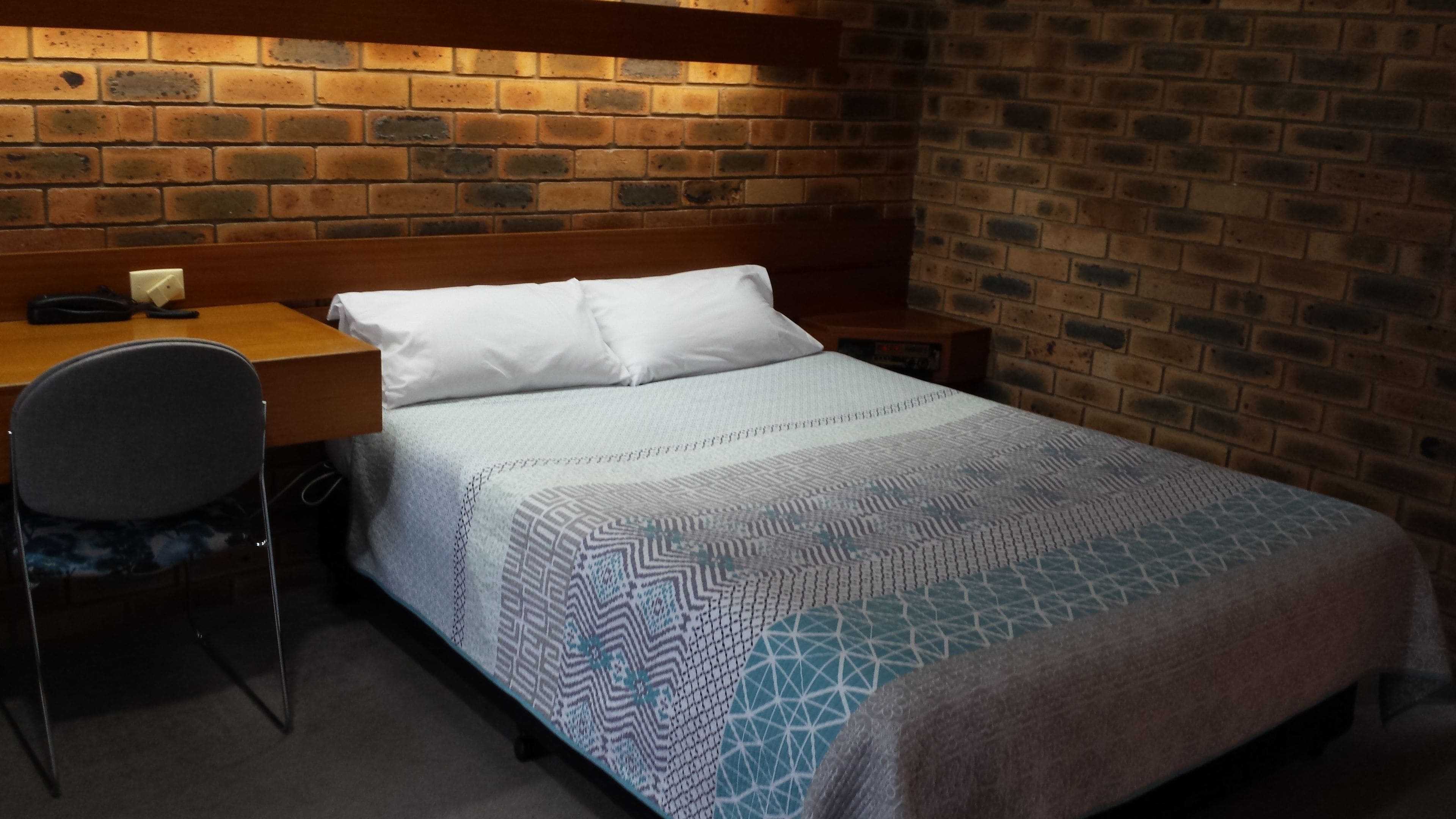 Una cama individual con una colcha estampada, un escritorio de madera y una silla en una habitación con paredes de ladrillo.