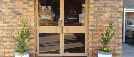 A brick building with a double door entrance, a visible emergency exit sign, and two potted plants on either side.