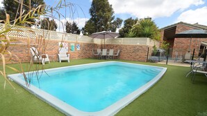 Una piscina rectangular con borde blanco, rodeada de césped artificial. Se ven sillas de descanso y una mesa con sombrilla al fondo.