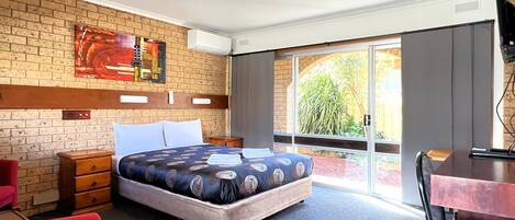 A hotel room with a bed, a desk, a chair, a TV, and a window with a view of greenery.