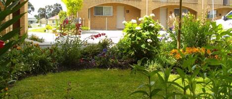 Um prédio de apartamentos com dois andares, fachada de tijolos, várias varandas e um jardim bem cuidado com diversas plantas e flores.