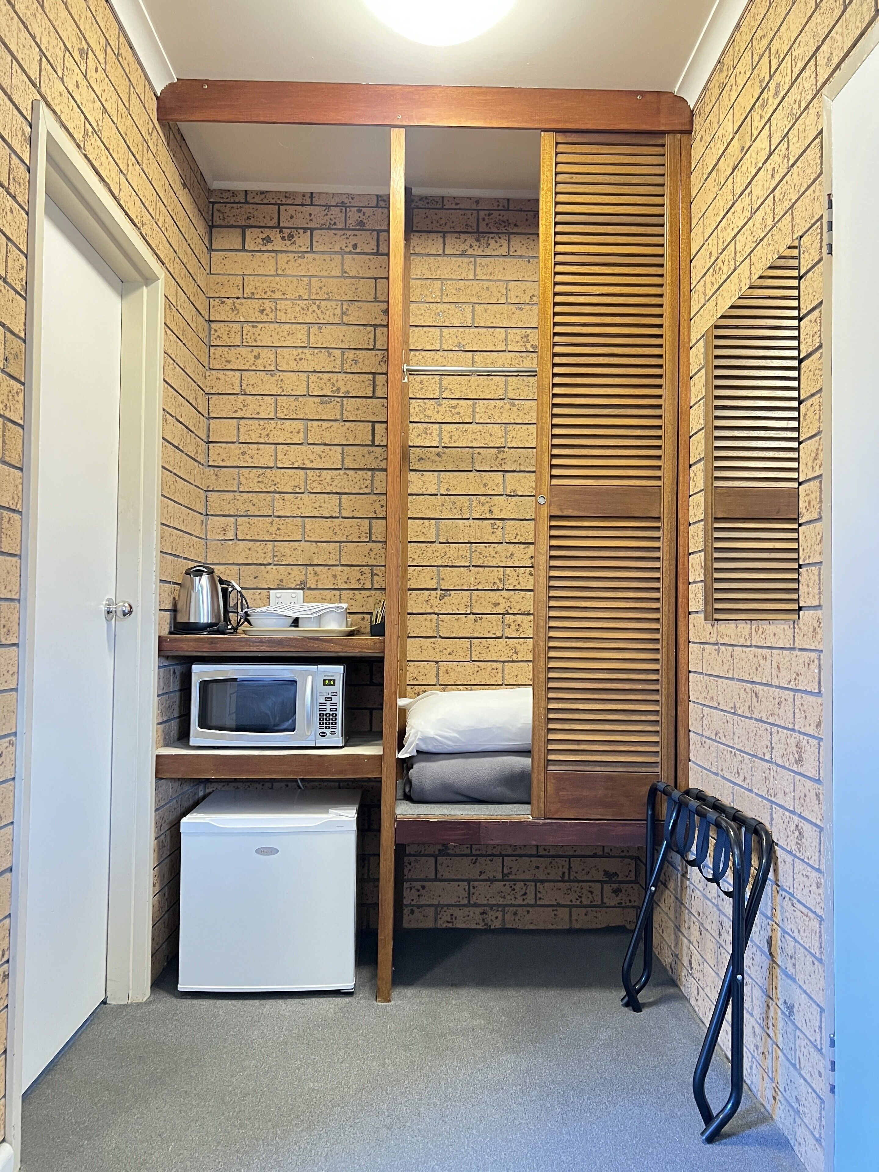 Habitación compacta con pared de ladrillo, estructura de cama de madera, microondas, refrigerador pequeño y una puerta.